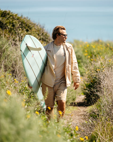 Man walking on trail near the ocean holding a surfboard, wearing the coach's vinyl jacket in tan over a tan t-shirt and tan corduroy shorts. 