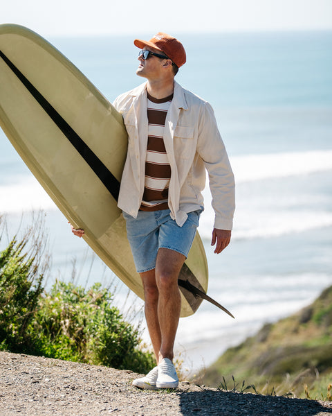 Man wearing milk colored Monterrey Shirt unbuttoned with t-shirt with horizontal tobacco stripes underneath. Man is holding a tan surfboard with black vertical stripe. He is wearing a rust colored corduroy hat and cardiff jean shorts with white shoes. Background showing green cliff and hill on right with blue ocean. 