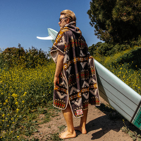 Kevin wearing the Pendleton x Birdwell Terry Poncho holding a mint green surfboard on a trail with yellow flowers and blue sky.