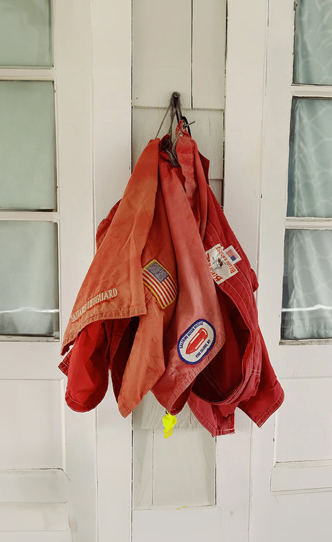 Three vintage red boardshorts hanging on a nail attached to a white wood slat wall and windows.