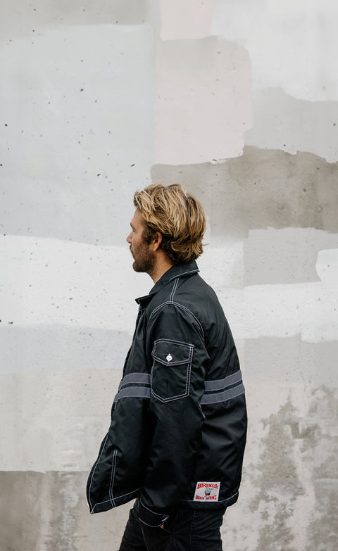 Side view of Kevin wearing black nylon jacket with slate stripes in front of cement wall background.