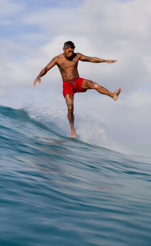 Toots wearing the red 300 nylon boardshorts while balancing on one leg with arms out on a surfboard while riding on a blue wave.