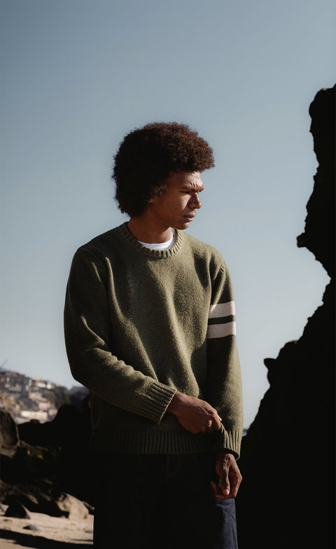 Obi wearing the comp stripe sweater in olive layered over white t-shirt and dark blue pants. Background depicts a rock formation on the right side and an ocean cliff side with homes with blue sky.