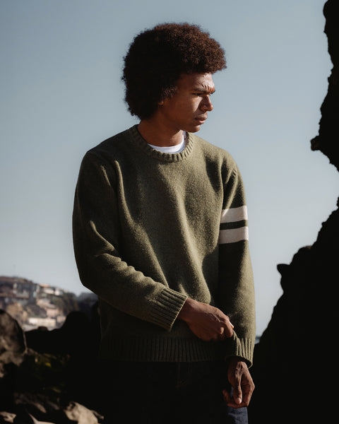 Obi wearing the comp stripe sweater in olive layered over white t-shirt and dark blue pants. Background depicts a rock formation on the right side and an ocean cliff side with homes with blue sky.