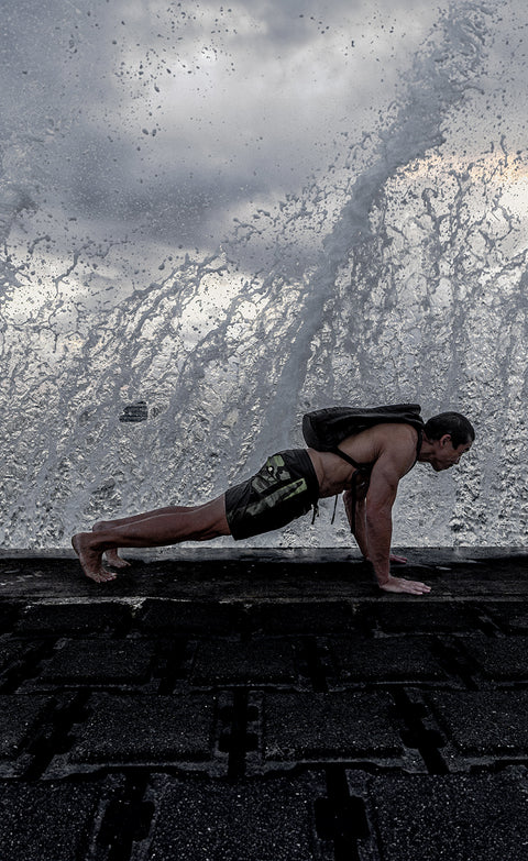 Bill Maeda doing pushups on the ground with a wave splashing into the air behind him. Wearing the GR1 backpack and 300 Boardshorts in Black and Camo.