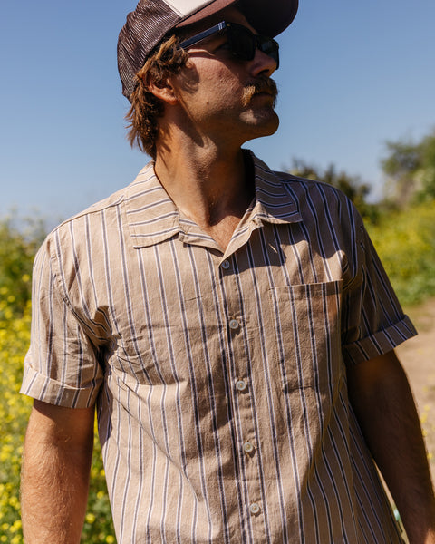 Man wearing Solana Shirt, a tan button up with vertical blue and white stripes with left chest pocket. Wearing sunglasses and brown mesh hat. Background showing green trail with blue sky.