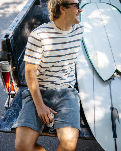 Man sitting on vintage black truck bed wearing horizontal striped t-shirt, federal blue corduroy shorts and dark sunglasses while holding can. Two white surfboards with thin vertical stripe laying in the bed of the truck.