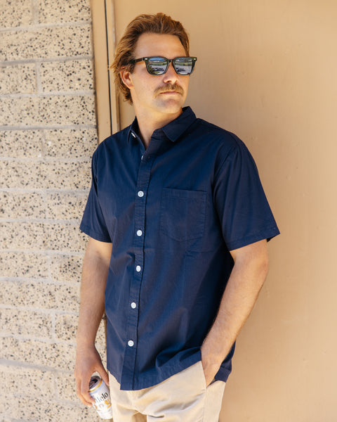 Man leaning against a tan brick wall and door wearing the dark blue Sandpiper button up shirt, tan shorts and, dark sunglasses while holding a drink in a can.