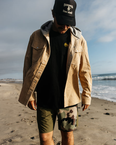 Man walking on the beach looking down. Wearing the Cambria tan corduroy jacket with grey hood over a black t-shirt. Wearing boardshorts with one camo leg and one army green leg and black foam hat with Birdwell logo.