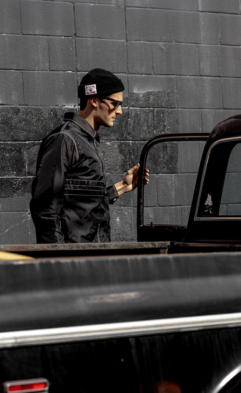 Eli wearing Comp Jacket 25 - Black/Black and black beanie while opening vintage black truck door. Black brick wall in background.