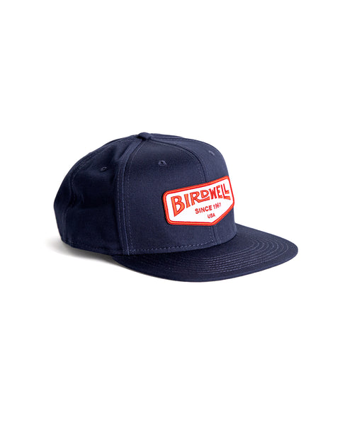 Navy snapback with bill pointing to the right. Patch in the center of the front of hat with embroidered Birdwell Logo in Red and Since 1961 USA below the logo. Product on white background.