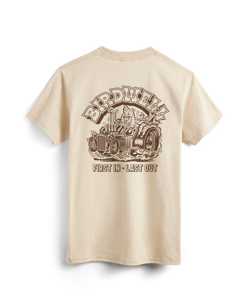 Back view of sand t-shirt with screen printed graphic in brown on the back of shirt. Screen print depicts Birdie in a vintage race car with large back wheels with smoke coming out. Birdwell is written above in an outlined Slab Serif font. 