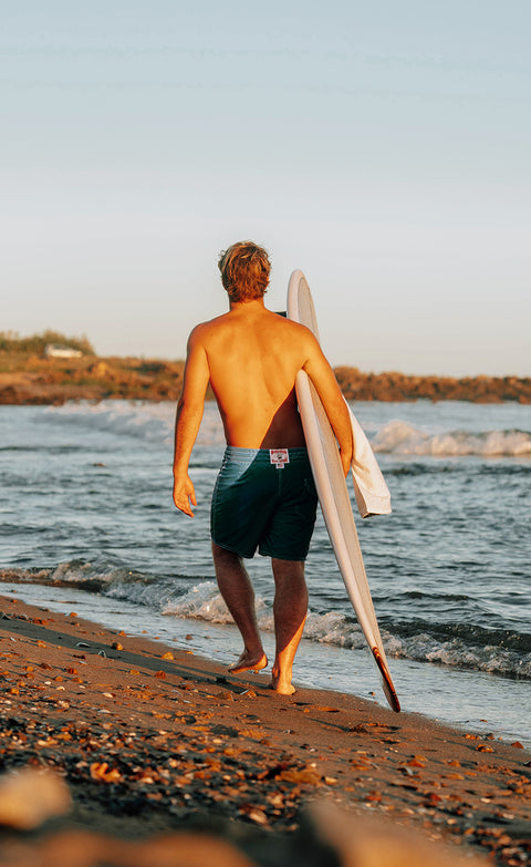 Kevin wearing the 300 boardshorts in dragonfly while holding a white surfboard in right arm. Beach in background.