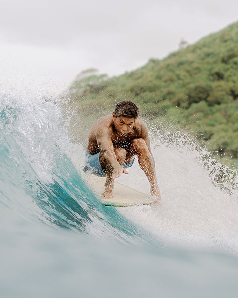 Toots wearing the 300 boardshort while surfing on teal blue wave. Island hill in background covered in green trees.