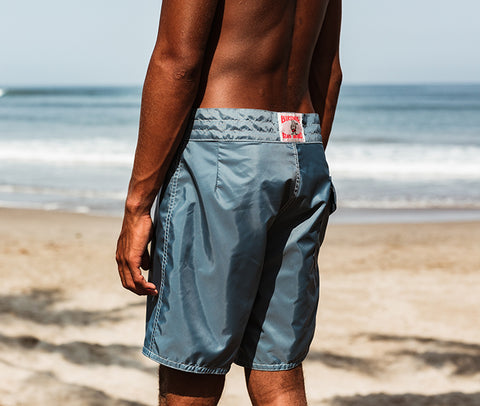 Surfer standing on the beach in federal blue boardshorts with shore in background.