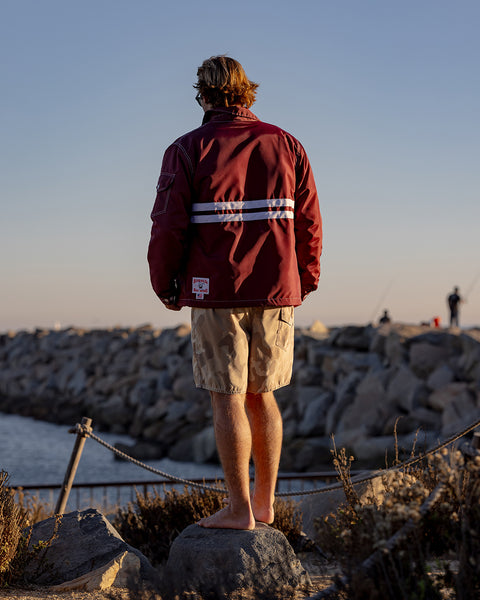 Kevin with his back towards the camera wearing a burgundy jacket with horizontal white stripes and desert camo boardshorts. Kevin is standing on a rock with jetty in the background.