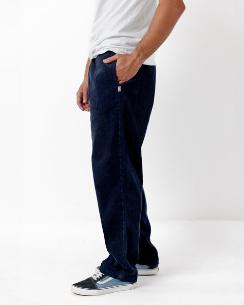 Model standing in front of white background wearing the Balboa Pants in navy with hand in pocket. Model is wearing white shirt and navy and light blue skating shoes.