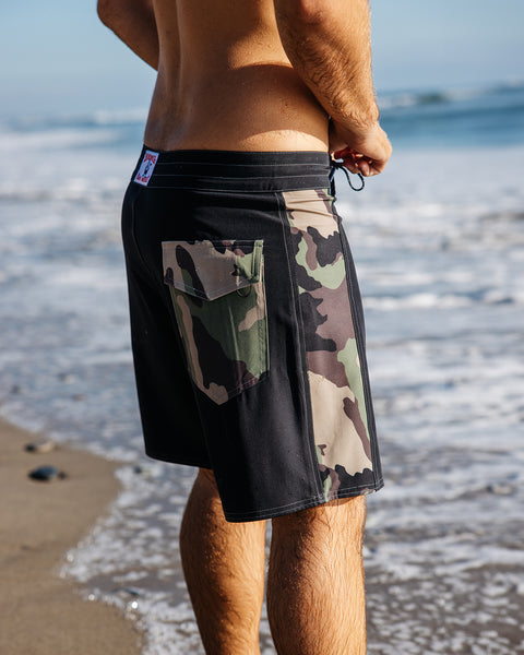 Model wearing the Black Camo Boardshorts while standing at the edge of the shore while tying the black drawcord. Pocket has a camo contrast fabric and camo panel shown on right leg.