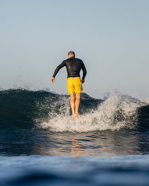 Christian Wach standing on a surfboard facing the front of the wave with his back towards the camera. Christian is wearing yellow boardshorts and a black wetsuit top. Background shows blue ocean waves and clear light blue sky.