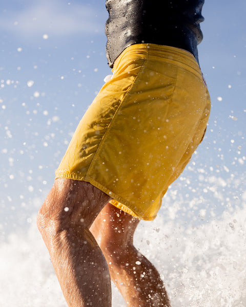 Close up shot of the yellow boardshorts with Christians right leg towards the camera. Black wetsuit top. Background shows splashes from the wave and bright blue sky.