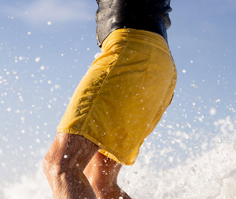 Surfer wearing yellow boardshorts and back wetsuit top with splashes from wave in background and blue sky