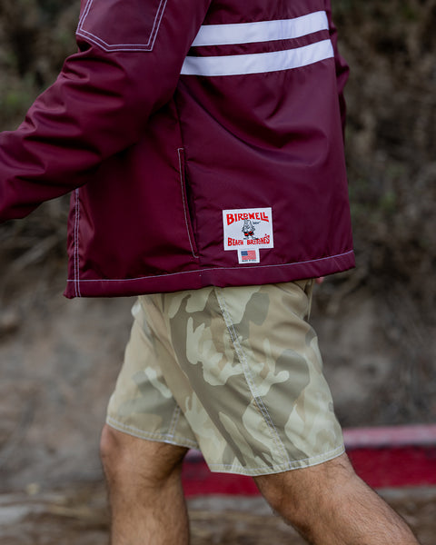 Model wearing the Comp Jacket 25 in Burgundy facing the left with two horizontal white stripes. Pocket and License plate label and made in usa label at the bottom left back of the jacket. Model wearing 300 Boardshorts in Desert Camo. Brown hill in background with red curb.