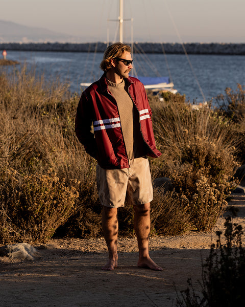 Kevin wearing the Comp Jacket 25 in Burgundy layered over the Comp Stripe Sweater in Oatmeal and 300 Boardshort in Desert Camo with brown bushes and harbor in background with docked boats.