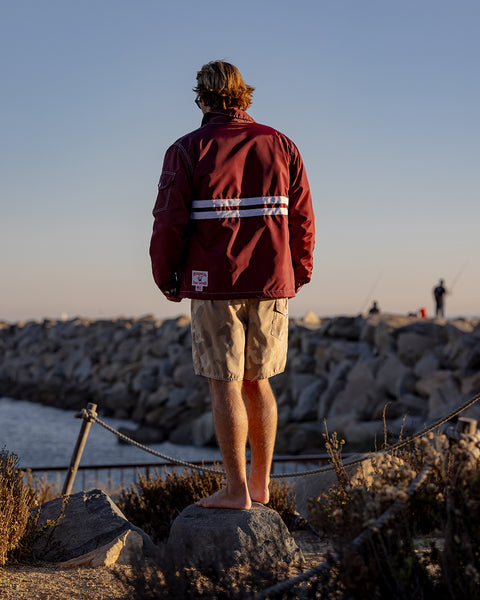 Kevin standing on a rock while facing his back towards the viewer wearing the Comp Jacket 25 in Burgundy with two horizontal white stripes and 300 Boardshort in Desert Camo with brown bushes and harbor in background with docked boats.