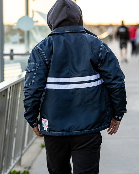 Model facing a metal railing on a pathway near the harbor wearing the Comp Jacket 25 in navy with two horizontal white stripes layered over a Grit Hoodie in Black.