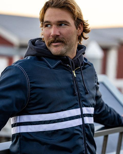 Kevin looking to the left with hands on a metal railing while wearing the Comp Jacket 25 in Navy with two horizontal white stripes layered over a Grit Hoodie in Black.