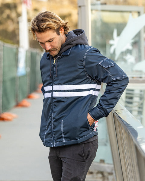 Kevin looking down wearing the Comp Jacket 25 in Navy layered over a Grit hoodie and black pants with hands in pockets. Metal railing to the right of model near harbor.