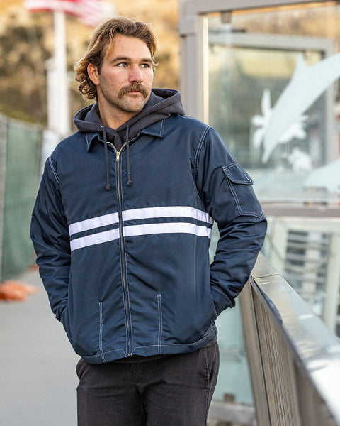 Kevin looking to the right wearing the Comp Jacket 25 in Navy layered over a Grit hoodie and black pants with hands in pockets.