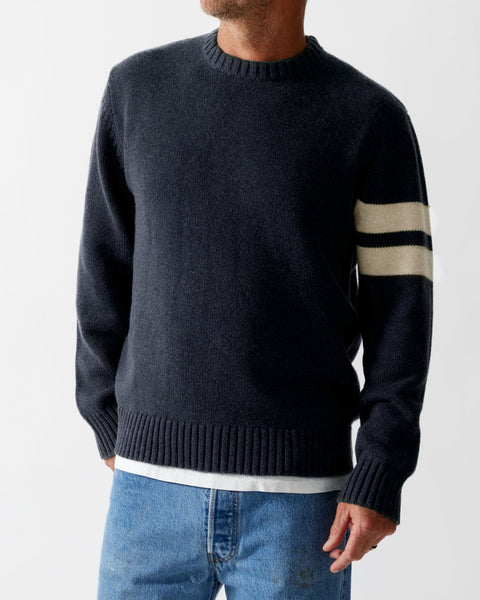 Model wearing Navy comp stripe sweater in navy and blue jeans with white shirt underneath. Grey background.