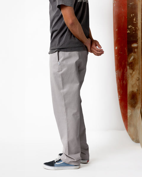 Model wearing dark grey shirt and Coronado Pants in Quarry with blue skating shoes. White background with red and dark brown sufboard standing in the upper right hand corner of the image.