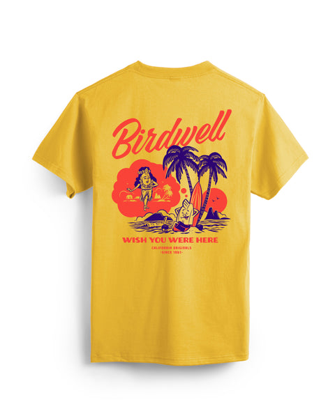 Flatlay of gold colored t-shirt on white background. Graphic depicting the birdie leaning against a surfboard under palm trees dreaming of a hula birdie with wish you were here written beneath in red and dark blue.