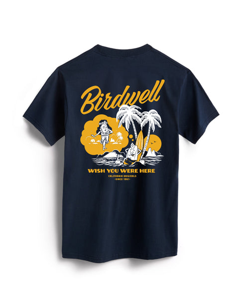 Flatlay of navy colored t-shirt on white background. Graphic depicting the birdie leaning against a surfboard under palm trees dreaming of a hula birdie with wish you were here written beneath in white and yellow.