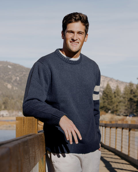 Bryce wearing the comp stripe sweater in navy one arm leaned against a wooden rail and the other hand in pocket. Blue sky and hills in background. Khaki pants. Wooden bridge in background.