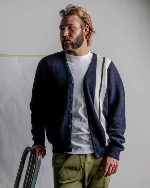 Lifestyle image of Kevin wearing the Comp Stripe Cardigan in navy layered over white t-shirt wearing army green coronado pants. He is leaning against a chair with white wall in background with white paper taped with tan painters tape to the wall.