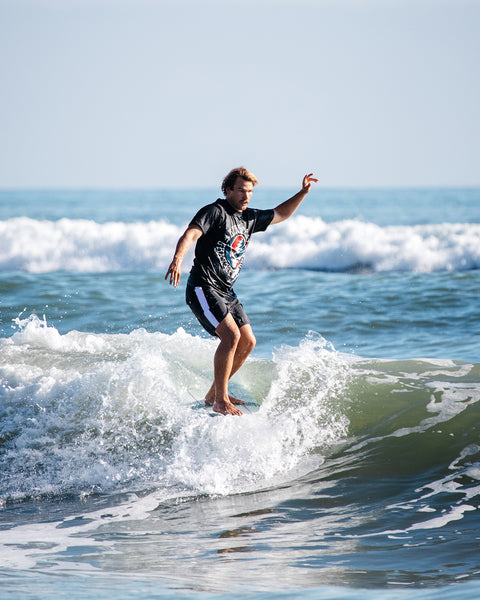 Kevin Skvarna surfing on the front edge of the board while wearing the Grateful Dead t-shirt and boardshorts. Background shows blue ocean waves and blue sky. 