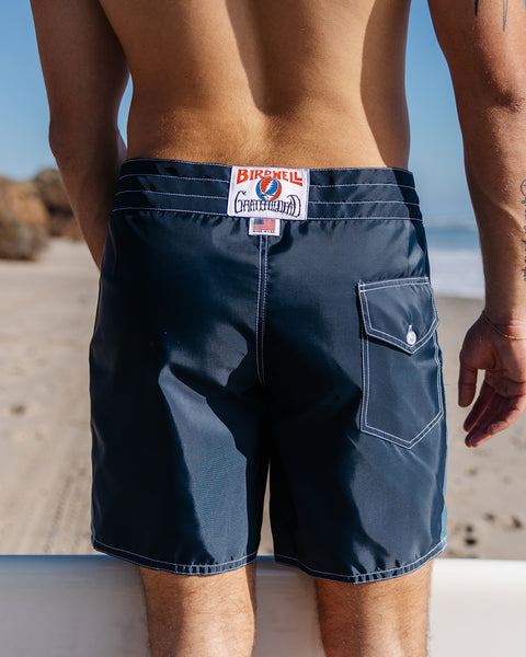 Surfer wearing navy boardshorts, image showing back of shorts. White label on waistband with Birdwell Grateful Dead collaboration logo, made in usa logo with american flag. Pocket on right side with white button. Beach in background with sand and ocean in the distance.