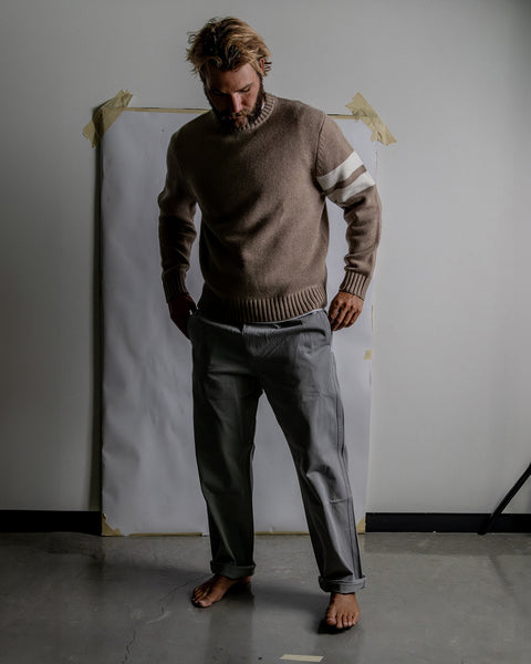 Kevin wearing the Comp Stripe Sweater in Oatmeal and Coronado Pant in Quarry while standing barefoot on a cement floor. White background with white paper taped to wall with painters tape.