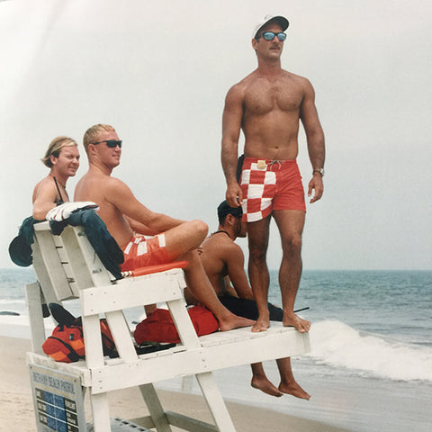 Bethany Beach Patrol Lifeguards in red checkered boardshorts while sitting on lifeguard stand at the beach.