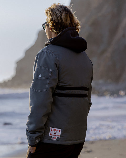 Kevin looking towards the ocean with back facing the camera with hands in pockets wearing the Quilt Lined Comp Jacket in Slate layered over a black Grit hoodie wearing black pants. Background is shore line with cliffs with tide rolling in and light blue sky.