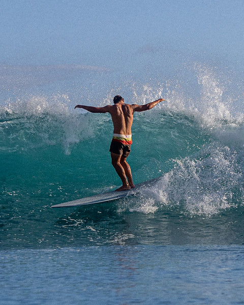 Kevin Skvarna surfing while facing a wave with his arms up in a T formation while wearing the Horizon Boardshorts with horizontal stripes in yellow, paprika and tobacco with blue wave in background.