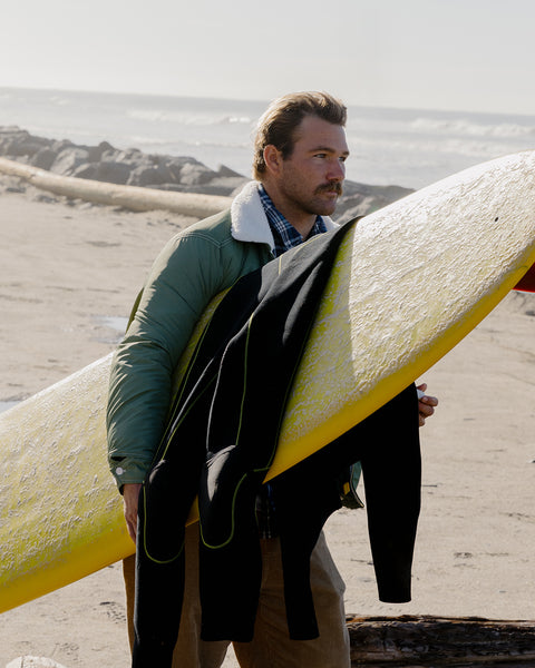 Kevin Skvarna wearing the Miramar Jacket in Olive over the Stinson Button Up Shirt in Navy while holding a yellow surfboard with black wetsuit draped over it. Background is a sandy beach with waves.