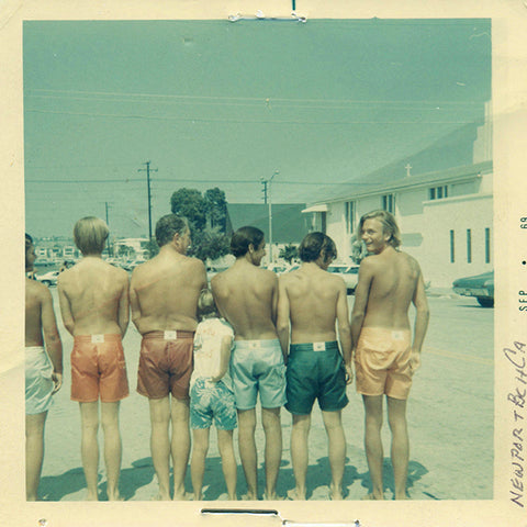 Vintage shot of 7 guys standing with their back towards the camera in different colored boarshorts.