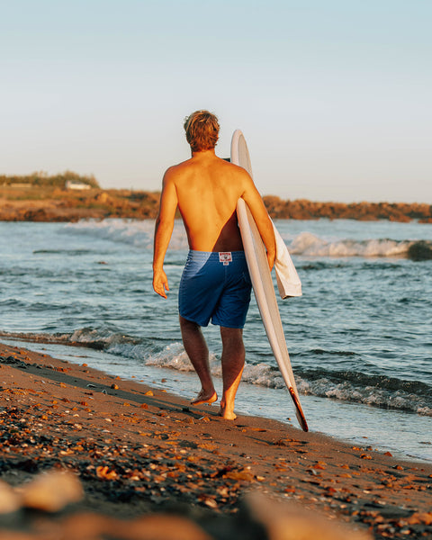 Kevin Skvarna wearing the Nylon 300 Boardshorts in Royal Blue white walking on the beach with calm waves rolling in. Background with blue sky and rocky shore.