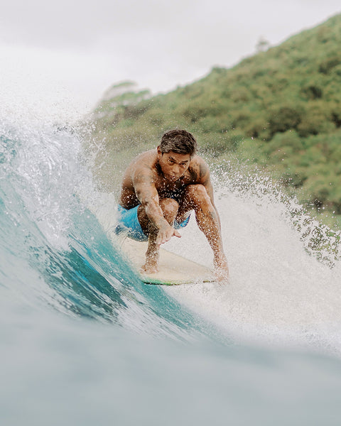 Toots wearing the light blue SurfNyl™ 300 Boardshorts in Light Blue while surfing on the nose of a board. Water splashing from the wave throughout the image with green island hill in background.