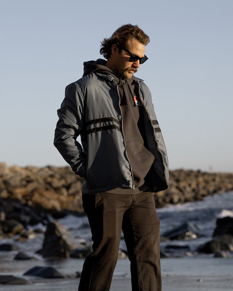 Kevin looking down with hands in jacket pockets wearing the Quilt Lined Comp Jacket in Slate layered over a black Grit hoodie wearing black pants. Background is shore line with rocks with tide rolling in with blue sky.