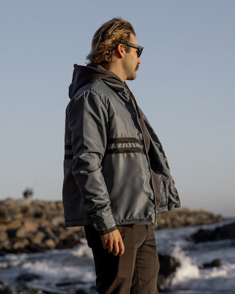 Kevin looking right towards the ocean wearing the Quilt Lined Comp Jacket in Slate layered over a black Grit hoodie wearing black pants. Background is shore line with rock ocean barrier with tide rolling in with blue sky.